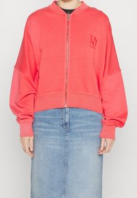 Sudadera con cremallera color coral, con cuello alto, mangas oversized, puños acanalados y un parche en la parte frontal. Lleva puesta sobre una falda de mezclilla. Textura de tela suave.