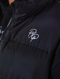 Chaqueta negra con cremallera frontal, con un panel superior texturizado y un panel inferior liso, adornada con un logotipo bordado en blanco.