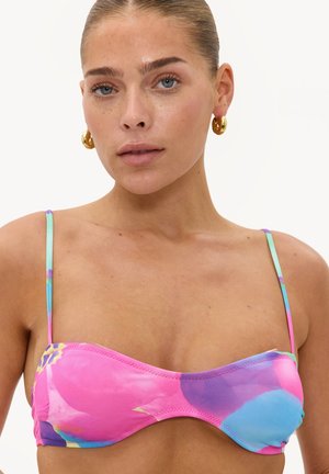 Femme aux cheveux plaqués en arrière portant un haut de bikini coloré rose, bleu et violet et des boucles d'oreilles créoles dorées sur un fond blanc uni.