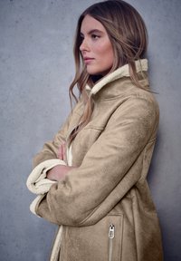 Chaqueta de pelaje sintético beige con un collar alto y cierre de cremallera. Presenta una textura suave y un forro interior de contraste, todo ello sobre un fondo gris.