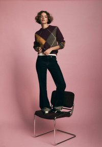 Maglione con un motivo a rombi in bordeaux, giallo e grigio, abbinato a pantaloni flare in denim scuro. Il modello è in piedi su una sedia marrone contro uno sfondo rosa.