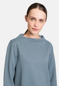 Bluse mit blauem Muster und strukturiertem Wabenmuster, mit langen Ärmeln und rundem Ausschnitt. Das Material wirkt leicht und atmungsaktiv.