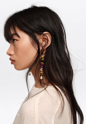 Joven con cabello largo y oscuro, llevando un pendiente largo y colgante hecho de cuentas multicolores y una blusa color crema, mirando hacia la izquierda.