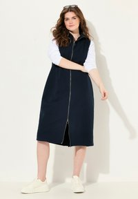 Abito blu navy con zip, colletto alto, tessuto strutturato, lunghezza al ginocchio, dotato di una zip frontale e abbinato a sneaker bianche.