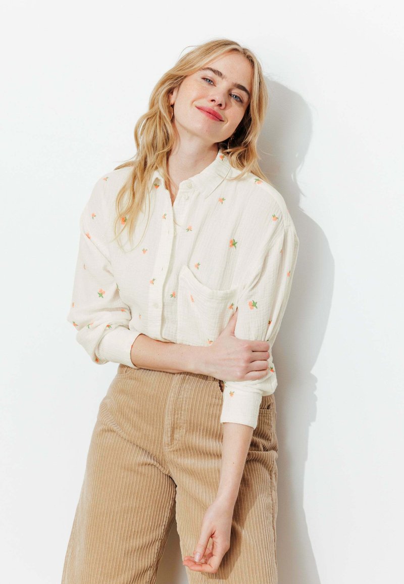 Jeune femme aux longs cheveux blonds portant une chemise blanche à fleurs à boutons et un pantalon en velours côtelé beige, debout sur un fond blanc.