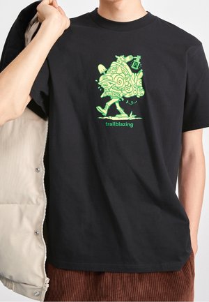 Osoba ubrana w czarny t-shirt z zielonym, kreskówkowym wędrowcem niosącym duży plecak oraz napisem „trailblazing” poniżej.