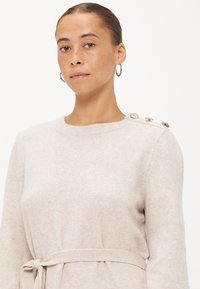 Lichtbeige trui van zachte stof, met een ronde halslijn, lange mouwen en knopenaccenten op de schouder. Inclusief een gestrikt tailleband.