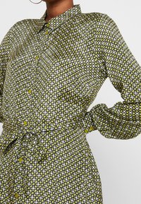 Green patterned shirt dress with a collared neckline, button-down front, and cinched waist. Features long sleeves and yellow button accents.