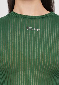 Zelený žebrovaný top s vysokým výstřihem, průhlednou strukturou a vyšitým logem "Stine Goya" v růžové barvě na levé straně.