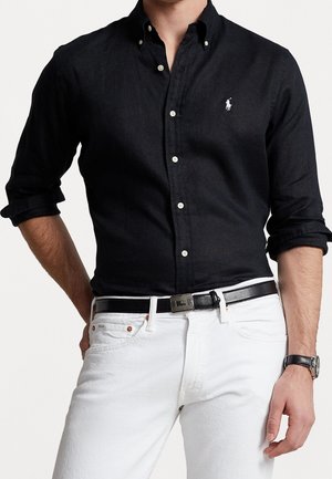Homme portant une chemise noire boutonnée avec manches retroussées, pantalon blanc, ceinture noire et montre noire au poignet, debout avec une main dans la poche.