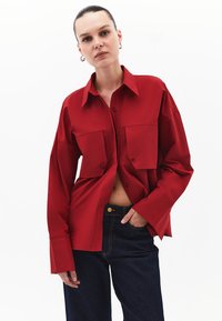 Camicia rossa con bottoni, due tasche frontali, vestibilità oversized, maniche lunghe e colletto a punta. Abbinata a jeans di denim scuro.