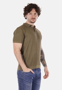 Polo shirt verde oliva con righe verticali testurizzate, maniche corte e colletto con bottoni. Indossato con jeans blu, mostrando tatuaggi sull'braccio.