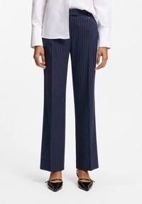 Pantalon rayé bleu marine avec une coupe ajustée, jambes larges et tissu lisse. Associé à des chaussures noires à bouts pointus avec des éléments décoratifs.