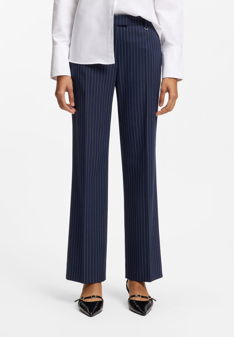 Pantalon rayé bleu marine avec une coupe ajustée, jambes larges et tissu lisse. Associé à des chaussures noires à bouts pointus avec des éléments décoratifs.
