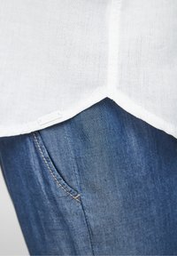 Vit linneskjorta med rundad fåll, som visar en blå denimtyg längst ner och en liten etikett. Ojämn fålldetalj och orange sömmar synliga.