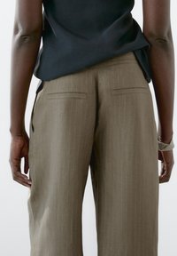 Hnědé pruhované kalhoty s volnějším střihem, vybavené dvěma zadními kapsami. Tkanina působí texturovaně a je doplněna jednoduchým černým topem.