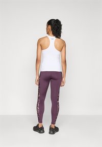 Vrouw die met haar rug naar de camera staat, gekleed in een witte racerback tanktop, paarse leggings met gestreepte zijkanten en zwarte sportschoenen.