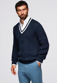 Granatowy sweter dzianinowy z dekoltem w serek, ozdobiony teksturowymi wzorami w warkocze oraz białymi detalami przy dekolcie, zestawiony z jasnoniebieskimi spodniami.