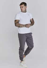 T-shirt bianco in cotone a maniche corte, pantaloni felpati grigi e sneakers bianche con lacci testurizzati. Presenta tatuaggi visibili sulle braccia.