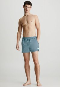 Costumi da bagno da uomo in tessuto teal, con vita elasticizzata, tasche laterali e un piccolo etichetta di accento. Tessuto liscio, lunghezza sopra il ginocchio.