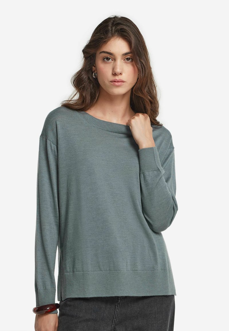 Kvinna med långt brunt hår, iförd en lös grågrön tröja och mörka byxor, håller tröjans krage med ena handen.