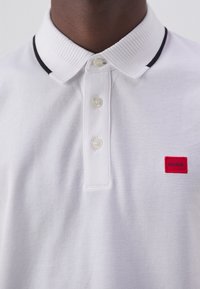 Polo shirt bianco realizzato in tessuto testurizzato, con colletto a costine con una striscia nera, tre bottoni e un'etichetta quadrata rossa con "HUGO."