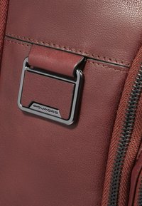 Detalle de bolso de cuero granate con hebilla de metal plateado, superficie texturizada, costuras contrastantes y una cremallera negra con acentos sintéticos.