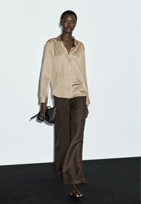 Camicia beige in raso con colletto e bottoni, abbinata a pantaloni larghi marroni. Il modello tiene una piccola borsa nera e indossa sandali aperti.