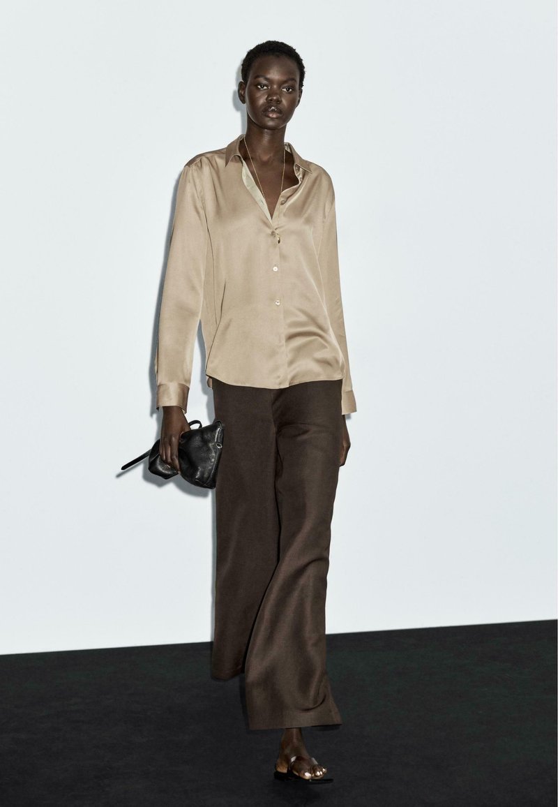 Camicia beige in raso con colletto e bottoni, abbinata a pantaloni larghi marroni. Il modello tiene una piccola borsa nera e indossa sandali aperti.