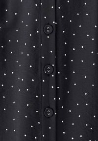 Czarna bluzka z tkaniny w białe kropki, z czterema okrągłymi guzikami z przodu i gładką fakturą.