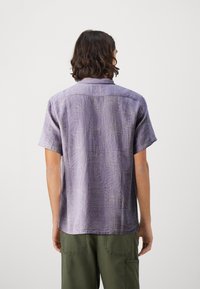Camisa morada de manga corta con un patrón texturizado. Tejido ligero con sutiles diseños geométricos y cuello con botones. Pantalones verdes debajo.