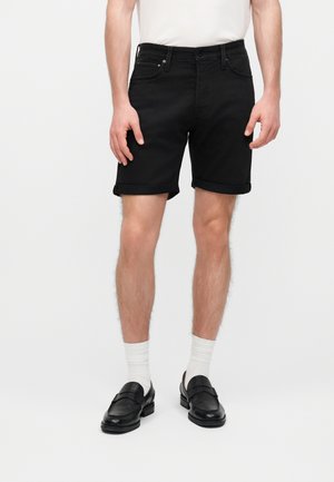 Mannlig modell iført svarte jeansshorts med oppbrett, hvite crew-sokker og svarte lær-loafers, stående foran en ensfarget hvit bakgrunn.