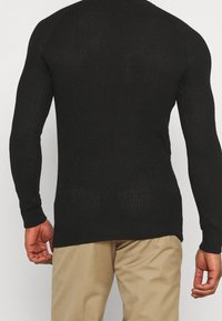 Svart stickad tröja med långa ärmar, ribbad textur och rak fåll, kombinerad med beige byxor. Bakifrån framhävs passformen och designen.