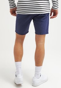 Námořnické modré šortky s jemně texturovanou tkaninou, ohrnuté lemování, nošené s bílými ponožkami a bílými teniskami. Viditelná je pruhovaná košile s dlouhým rukávem.