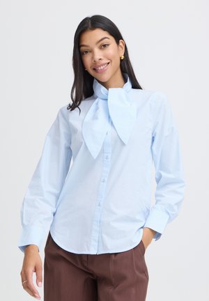 Mujer con cabello largo y oscuro, que lleva pendientes dorados y una blusa azul claro con un gran lazo en el cuello, sonriendo con una mano en el bolsillo de unos pantalones marrones.