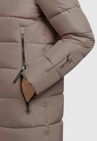 Giacca a vento in taupe tenue con design trapuntato. Presenta una tasca con zip su manica, hardware nero e una texture liscia e lucida.