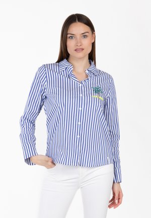 Frau trägt ein blau-weiß gestreiftes Hemd mit Knöpfen, das mit einer Palme und "SUMMER" bestickt ist, kombiniert mit weißen Hosen, stehend vor weißem Hintergrund.