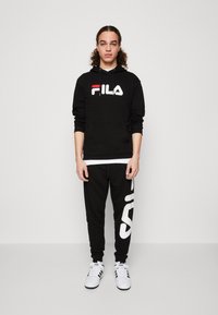 Felpa nera con logo Fila bianco, abbinata a pantaloni della tuta neri con una grande stampa bianca "Fila". Scarpe da ginnastica bianche completano l'outfit.