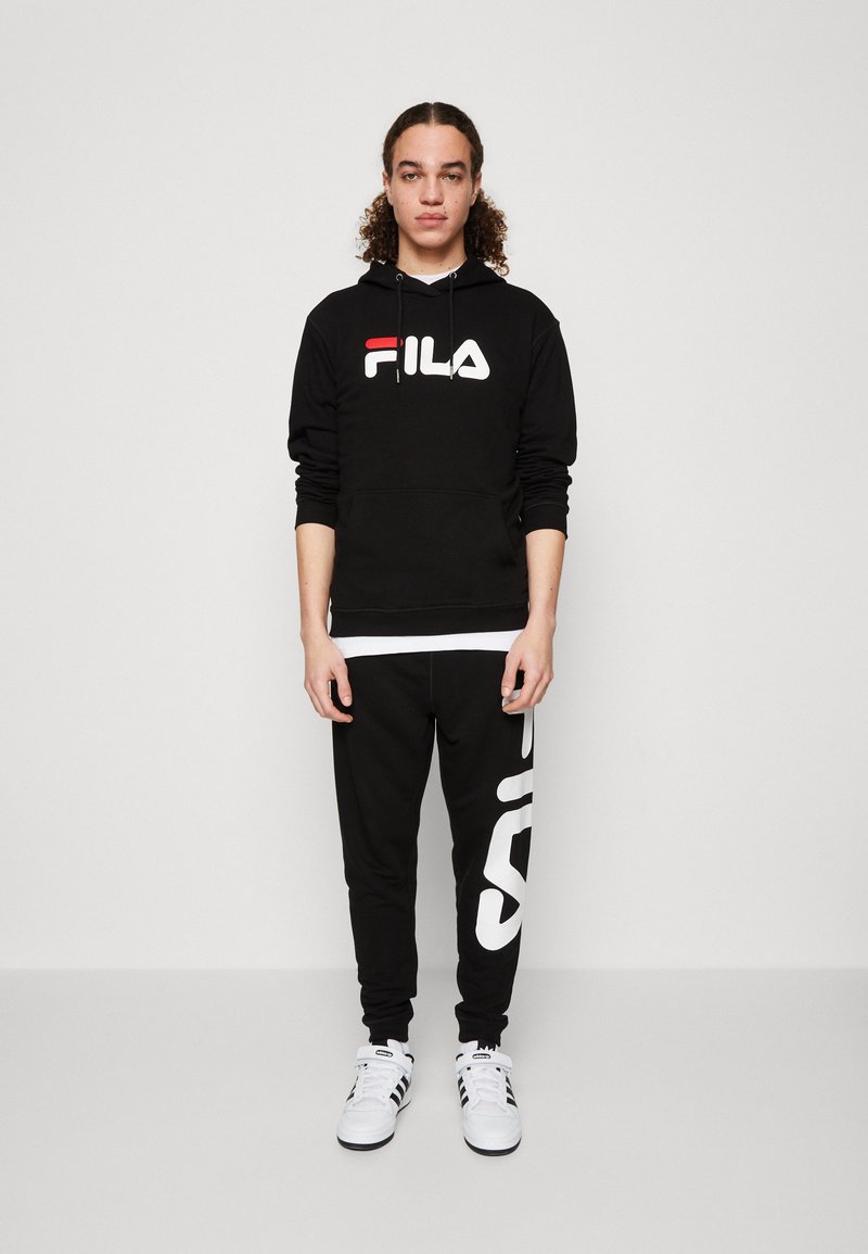 Felpa nera con logo Fila bianco, abbinata a pantaloni della tuta neri con una grande stampa bianca "Fila". Scarpe da ginnastica bianche completano l'outfit.