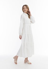 Hvid lang kjole med knappelukning foran, tieret design og lange ærmer. Bæret med beige hæle sandaler på en simpel baggrund.