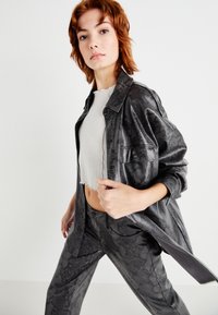 Schwarze Schlangenmustert-Lederjacke mit kragenlosem Ausschnitt, kombiniert mit einem strukturierten weißen Cropped-Top und passenden Hosen.