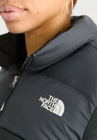 Černá péřová bunda s vysokým límcem, která má stříbrný zip a bílé logo "THE NORTH FACE" na levé straně. Texturovaná látka s viditelnými švy.