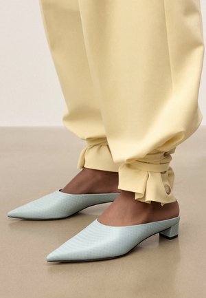 Füße in hellblauen spitzen Maultieren mit niedrigen Blockabsätzen, kombiniert mit blassgelben Hosen, die am Knöchel mit Knöpfen umgeschlagen und gebunden sind.