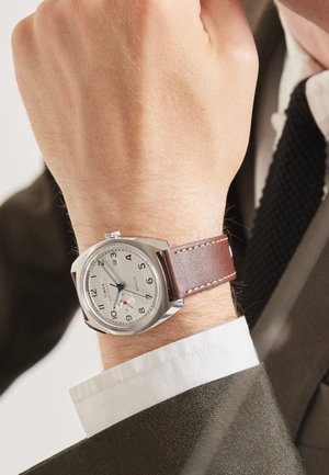 Montre argentée avec un bracelet en cuir brun, boîtier rond, cadran blanc, chiffres arabes noirs, aiguille des secondes bleue et fonction date à six heures.