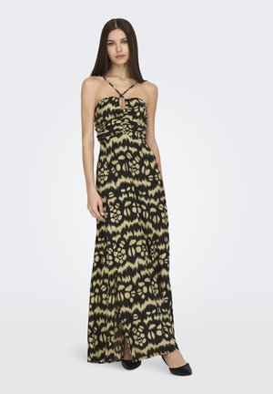 Mujer vestida con un vestido maxi de patrones negro y beige, con finas tiras entrecruzadas y una abertura en la parte delantera, de pie con una mano relajada a su lado.