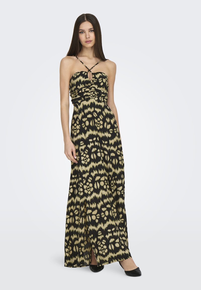 Mujer vestida con un vestido maxi de patrones negro y beige, con finas tiras entrecruzadas y una abertura en la parte delantera, de pie con una mano relajada a su lado.