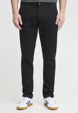 Black cotton chinos with a slim fit, featuring a classic button and zipper closure, side pockets, and a smooth texture. Paired with white sneakers.