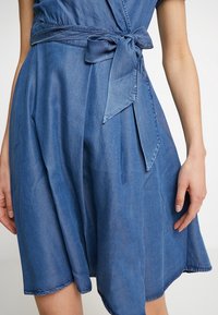 Denim-wrapped dress in medium blue, with a tied waist and short sleeves. The fabric is lightweight and has a soft, smooth texture.