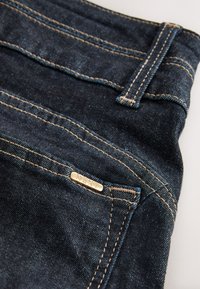 Jean en denim foncé avec des accessoires dorés, des coutures claires contrastantes et une poche avant dotée d'une étiquette métallique embossée du mot "NEXT".