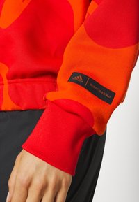 Sweat-shirt à motifs rouges et oranges avec poignets côtelés ; comporte des étiquettes Adidas et Marimekko ; tissu doux avec un design vibrant et audacieux.